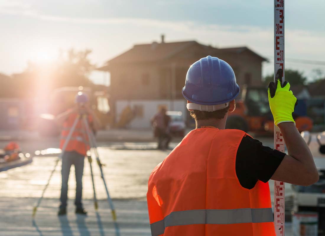 Land Surveyor Insurance - Level on a Tripod With Two Construction Workers in the Background of a Construction Site at Sunset
